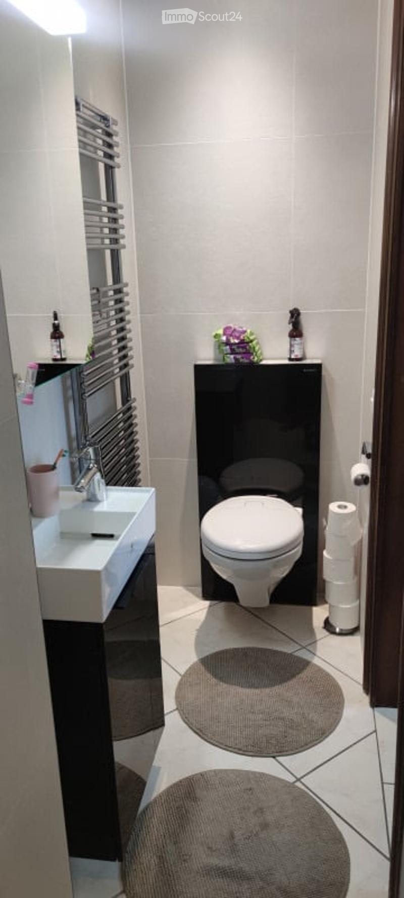 Modern bathroom with white tiles, a white sink, a toilet, a towel rack, and a small round rug on the floor.