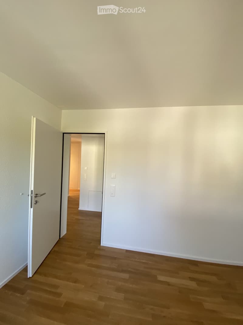 Empty hallway with light-colored walls, wooden flooring, and a partially open door leading to another room.