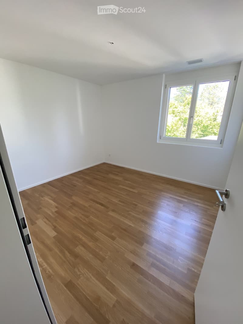 Empty room with light wooden flooring and white walls, featuring a large window with white frames and a door handle visible on the adjacent wall.