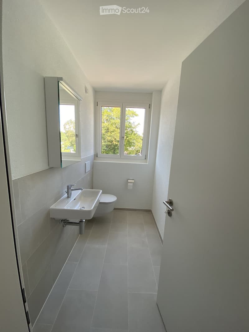 Modern bathroom with white tiles, a sink, toilet, and a large window offering a view of greenery.