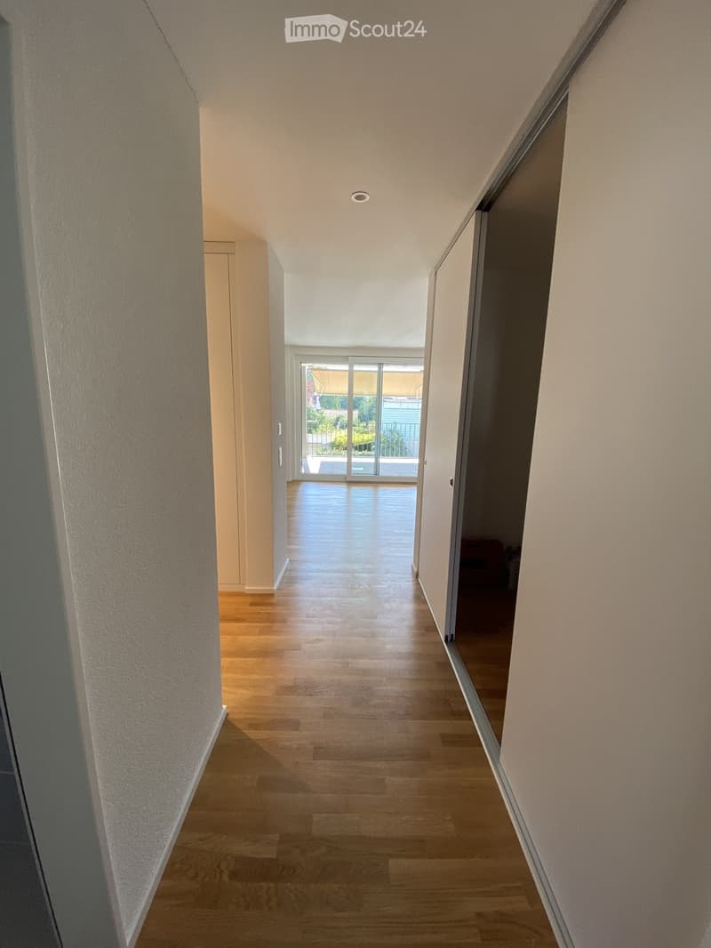 A modern hallway with wooden flooring, white walls, and recessed ceiling lighting. The hallway leads to a sliding glass door that opens to a balcony or outdoor area.