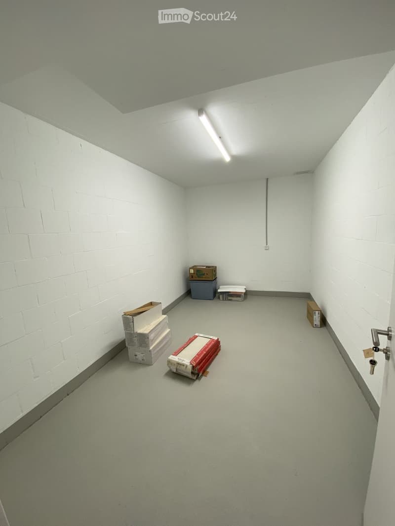 Empty room with white brick walls, grey floor, and fluorescent lighting. Several boxes and a small container are placed against the walls.
