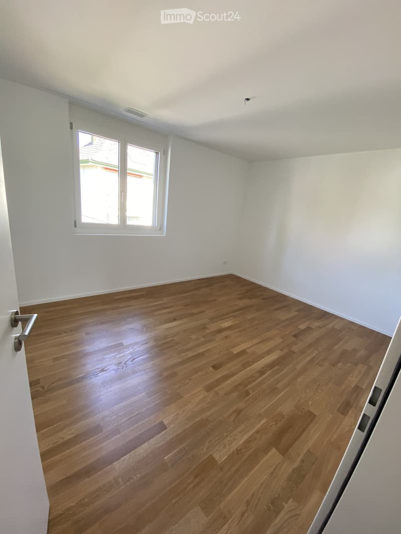 Empty room with light wooden flooring and white walls, featuring a large window with white frame and a door handle visible on the left side.