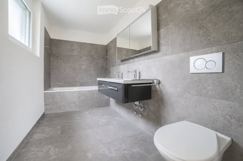 Modern bathroom with grey tiled walls and floor, double sink with chrome faucets, large mirror, and a toilet.