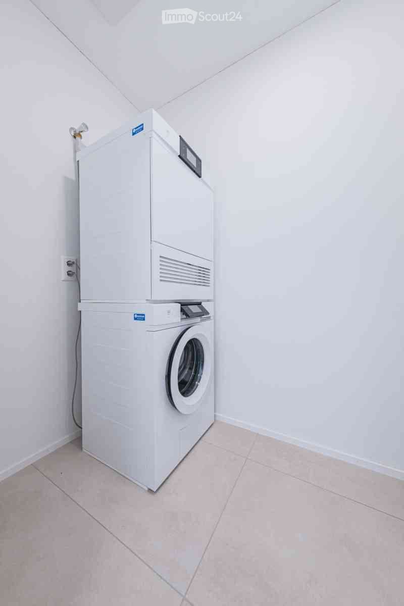 Empty room with white tiled floor and walls, featuring a stacked washer and dryer unit in the corner.