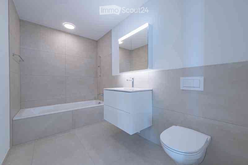 Modern bathroom with a bathtub, sink, toilet, and a large mirror with built-in lighting. The walls are tiled in neutral tones, and the floor is covered with light-colored tiles.