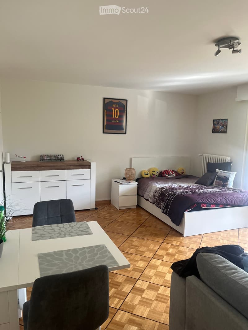 A modern, well-furnished room with a bed, sofa, table, and nightstand. The room features wooden flooring, white walls, and decorative items including a framed jersey and a small cabinet.