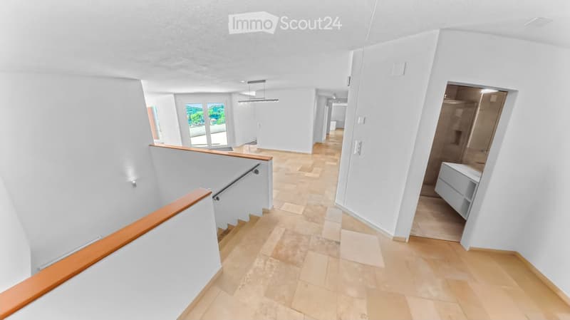 Bright, modern hallway with white walls, wooden flooring, and a staircase leading upwards. Features include recessed lighting, a wooden railing, and a view of a room with large windows and a balcony.