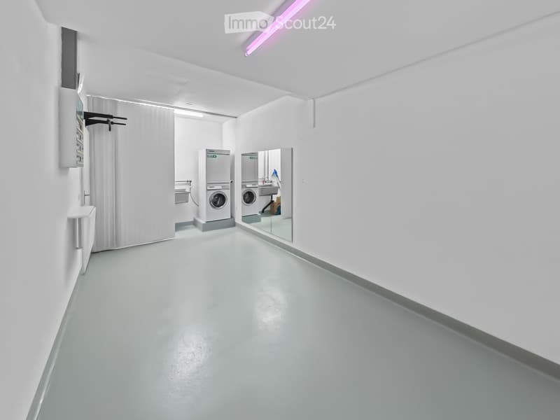 A clean, modern laundry room with white walls, a grey floor, and two stacked washing machines and dryers. A large mirror and a curtain are visible.