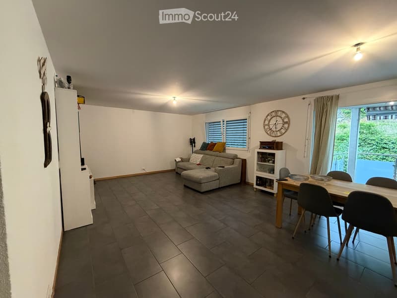 Open-plan living and dining area with a grey sofa, ottoman, dining table with chairs, and a small cabinet with glassware.