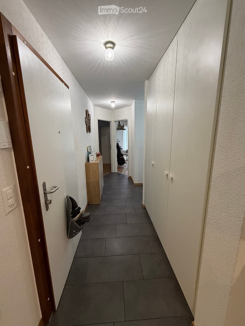 A narrow hallway with grey tiled flooring, white walls, and built-in white cabinets on one side. A small wooden cabinet with framed pictures and a shoe rack is visible on the left, with a door leading to another room at the end.