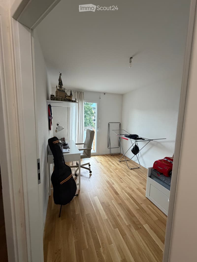 A small, well-lit room with wooden flooring, a desk with a chair, a guitar case, a lamp, a radiator, a curtained window, and a small bed with a toy on it.