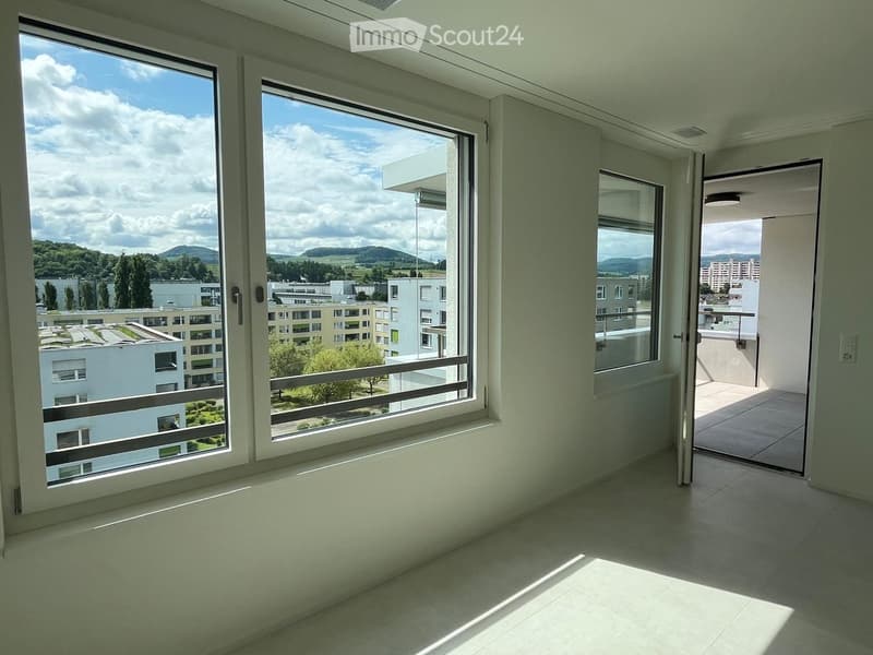 Empty room with large windows and a sliding door leading to a balcony, offering a view of the city and surrounding landscape.