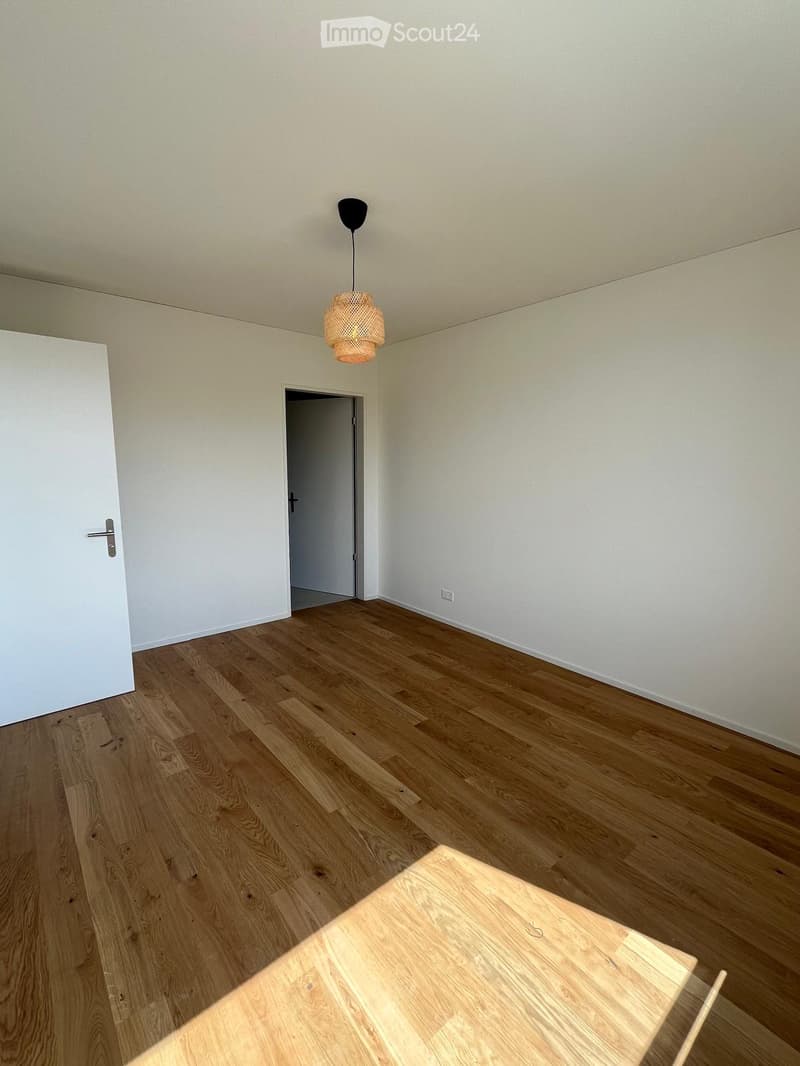 Empty room with light wooden flooring, white walls, and a single hanging light fixture. A small wooden table is partially visible in the foreground.