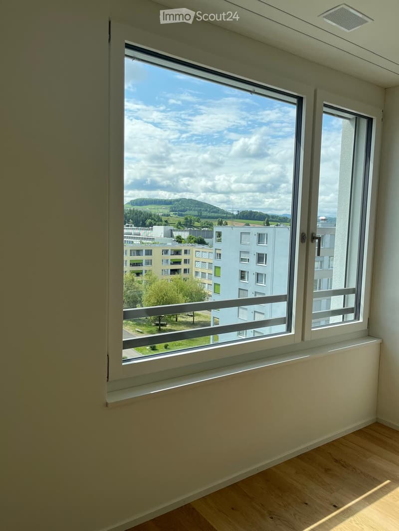 Empty room with wooden flooring, large window offering a view of the landscape and buildings, white walls, and a simple window sill.
