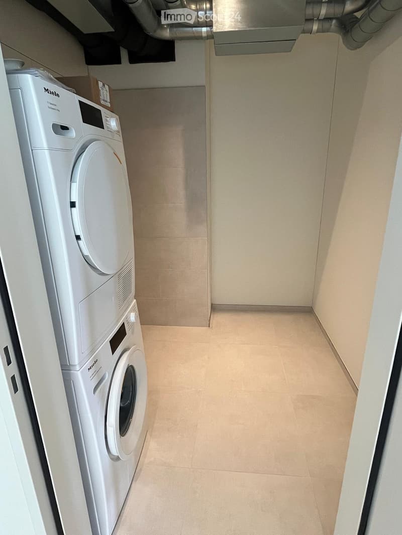 Narrow hallway with beige tiled floor and walls, featuring a stacked washing machine and dryer unit on the left side.