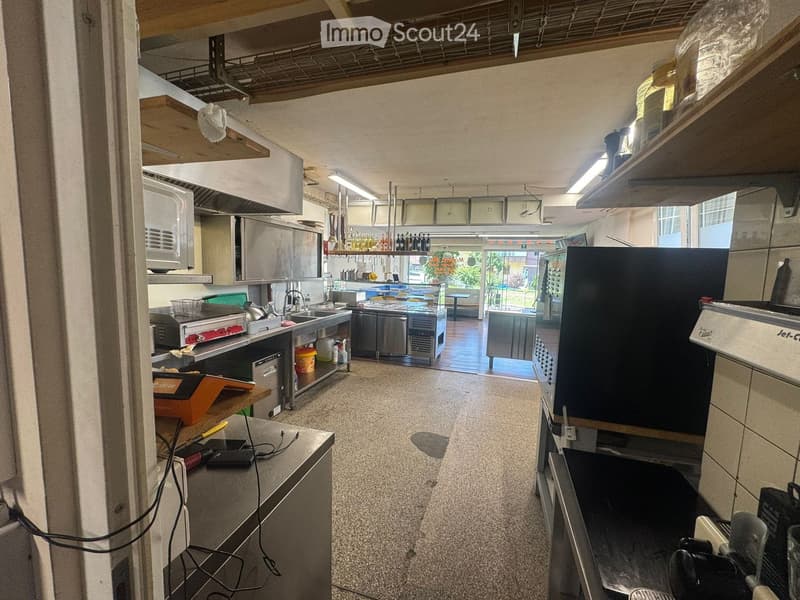 Commercial kitchen with stainless steel appliances, including a refrigerator, oven, sink, and counter. Shelves and cupboards are visible, along with various kitchen tools and equipment.