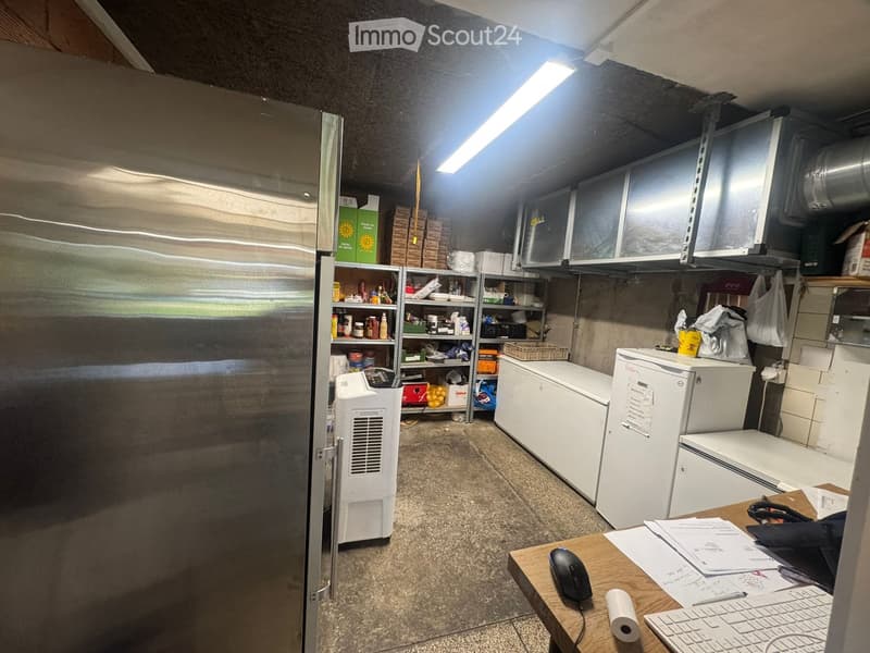 A small storage or utility room with a large stainless steel refrigerator, multiple freezers, shelves filled with supplies, a wooden table with a computer setup, and various boxes and storage items.