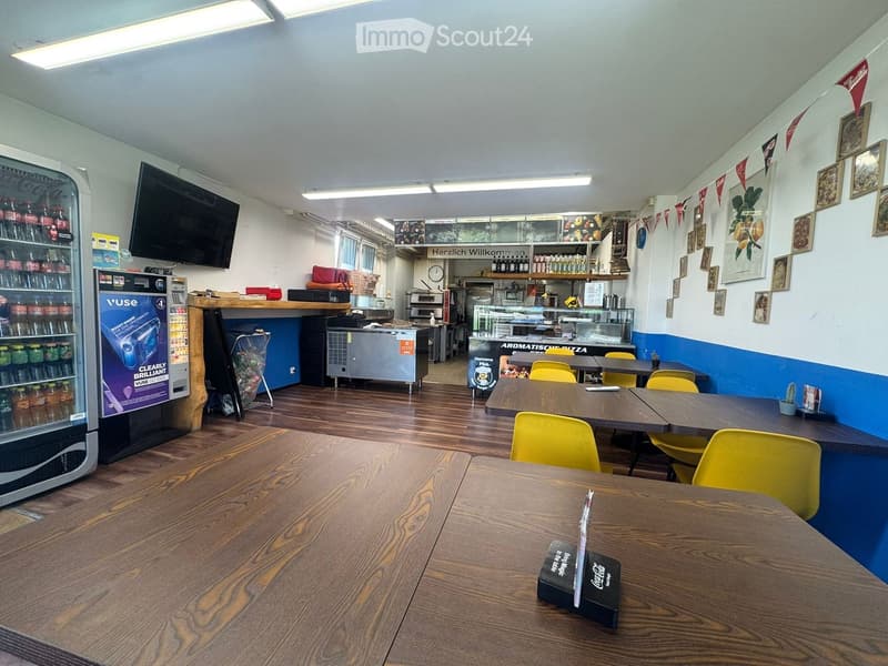 Restaurant interior with wooden tables, yellow chairs, a refrigerator, a screen, a counter, shelves, and picture frames on the walls.