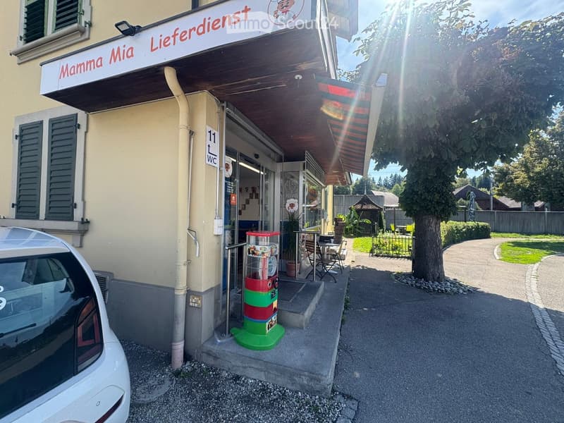Facade of a restaurant named "Mamma Mia Lieferdienst" with a signboard, entrance, vending machine, and outdoor seating area with tables and chairs.