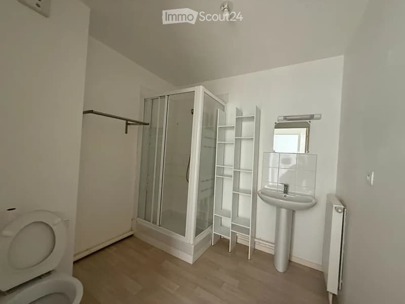 Modern bathroom with wooden flooring, white walls, a toilet, a sink with a mirror, a glass-enclosed shower stall, and a white shelf unit.