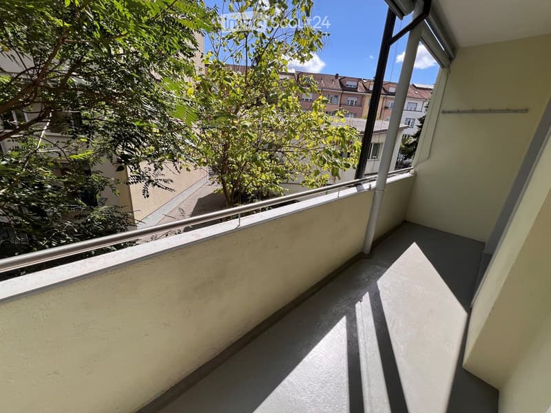 Balcony with metal railing, overlooking a tree-lined street and neighboring buildings.