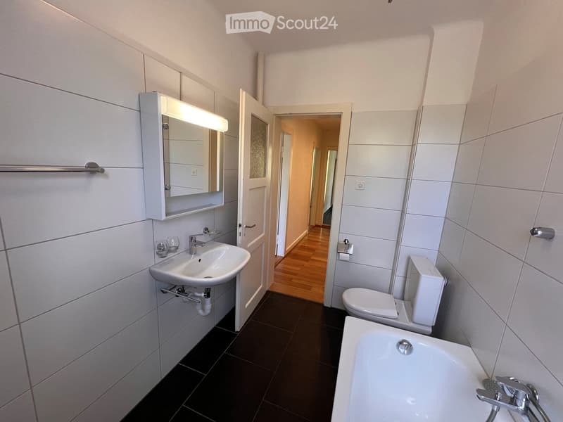 Modern bathroom with white tiles, dark floor, sink, mirror, bath, and toilet. The door leads to a hallway with wooden flooring.