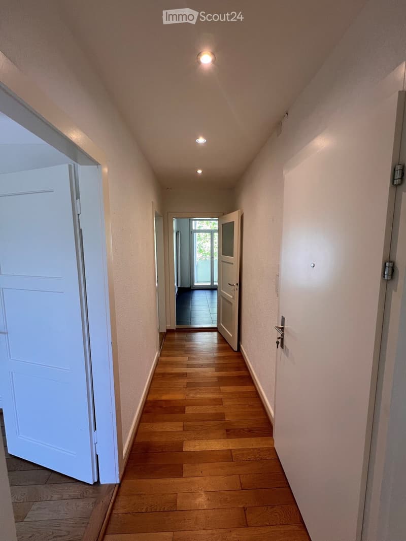 A narrow hallway with wooden flooring, white walls, and several doors leading to different rooms. The hallway is well-lit with recessed ceiling lights.