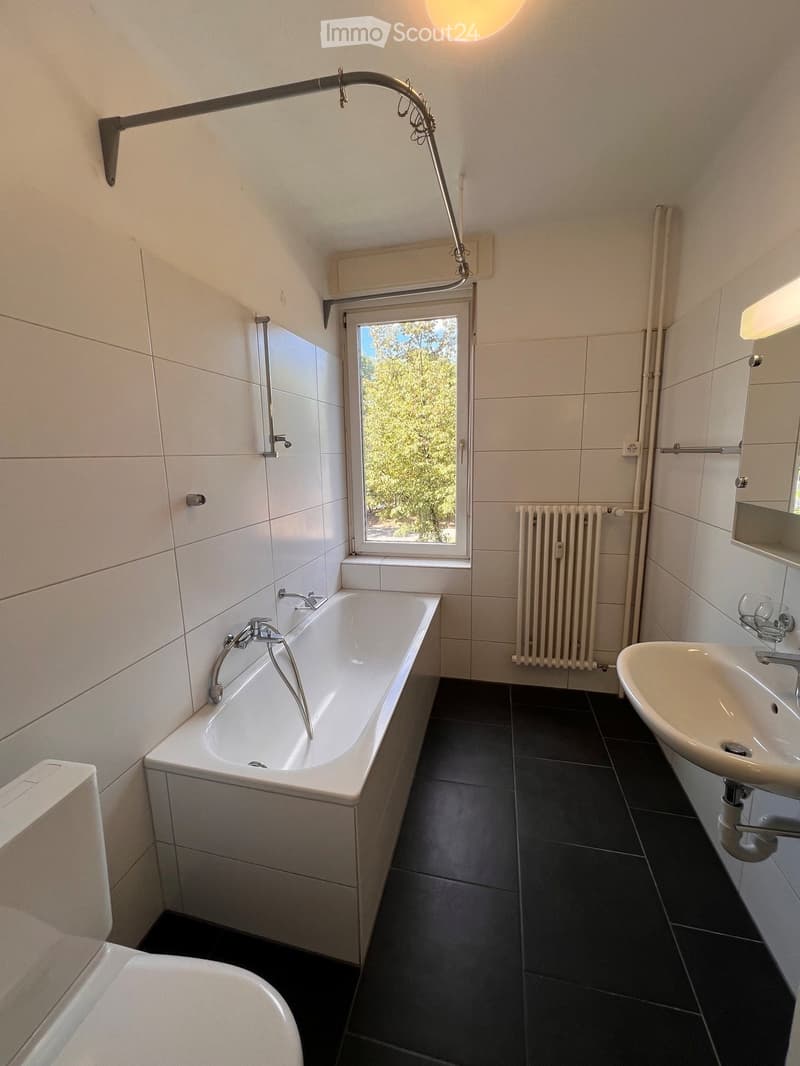 Modern bathroom with a bathtub, sink, radiator, and large window offering a view of greenery outside.