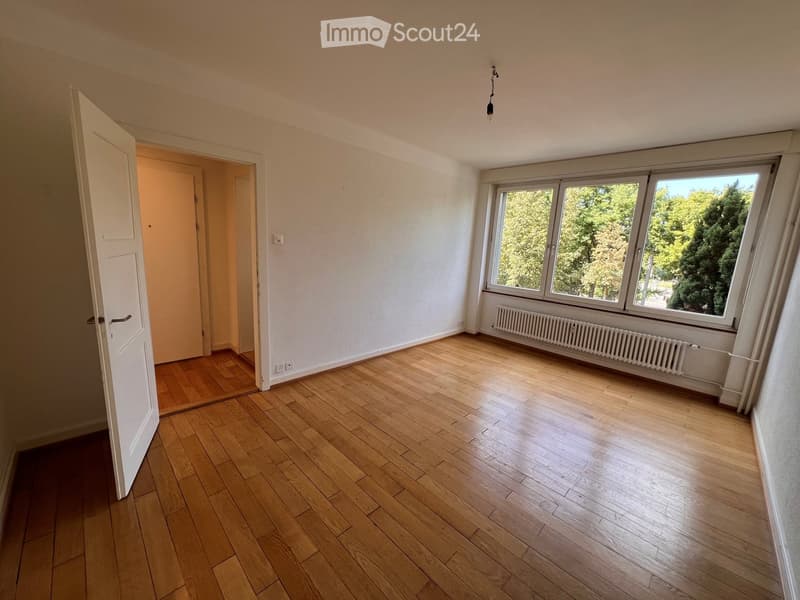 Empty room with wooden flooring, white walls, large windows with a view of greenery, and a radiator below the windows.