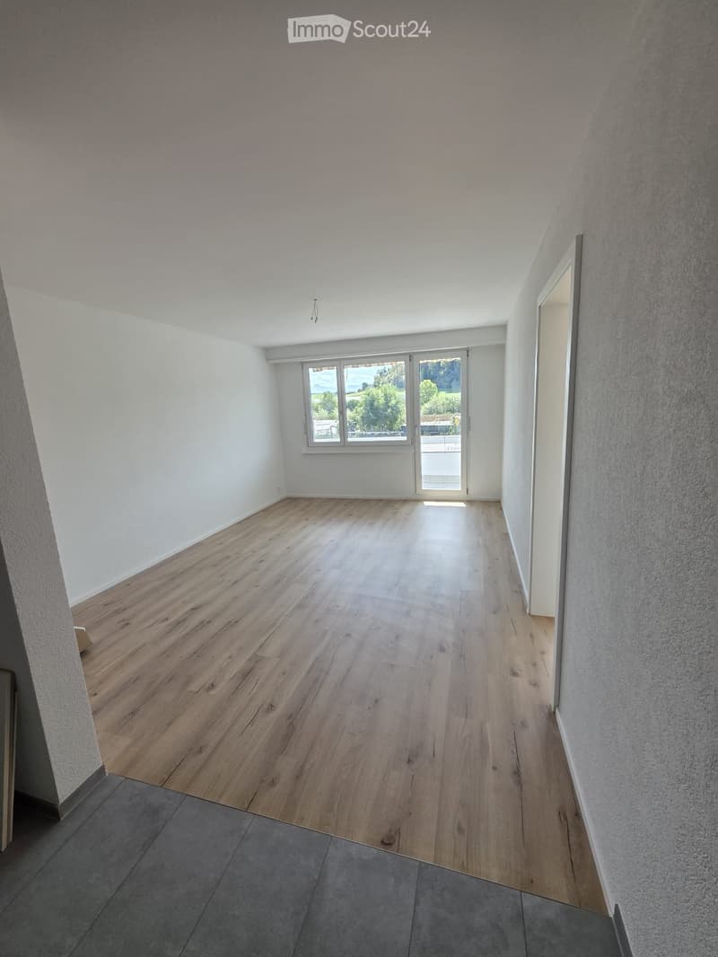 Empty room with wooden flooring, white walls, and large windows offering a view of the outdoors.