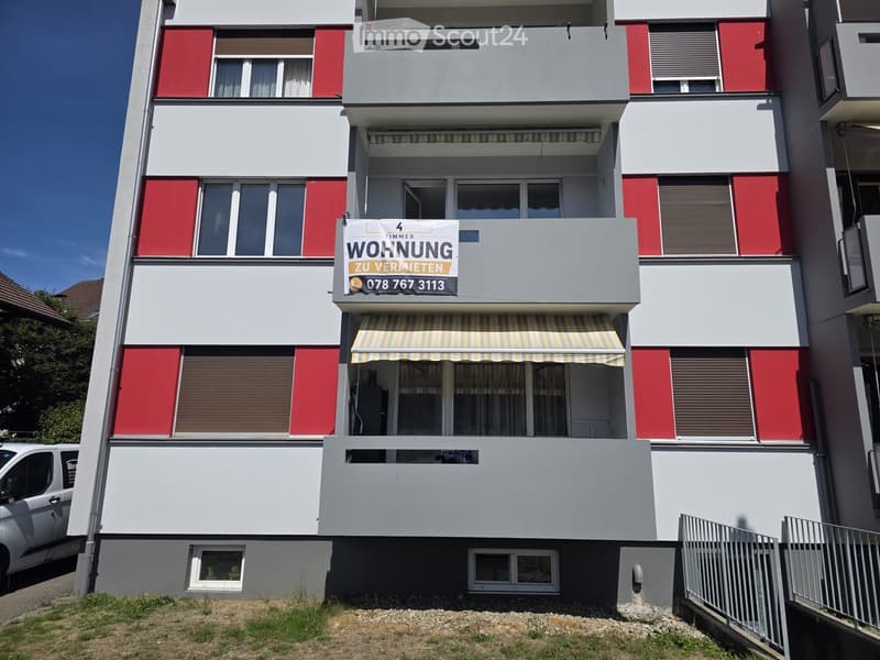 Four-story apartment building with red and white facade, multiple windows, balconies, and a rental sign on the third floor.