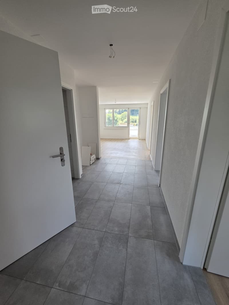 Empty hallway with tiled floor, white walls, and a ceiling light fixture. Doors lead to adjacent rooms, and a window at the end provides natural light.