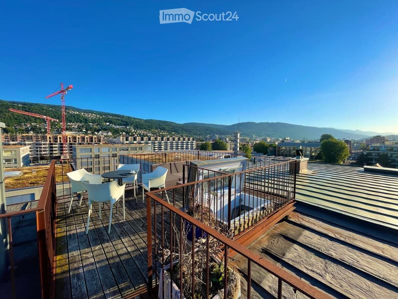 Roof terrace with wooden decking, metal railing, and a small seating area with a table and chairs, overlooking a cityscape with hills and construction cranes.