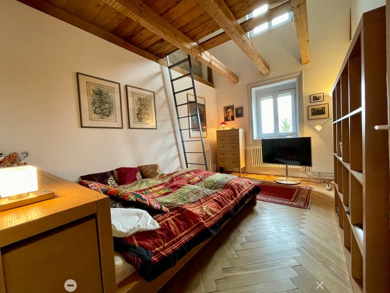 Cozy bedroom with wooden floors, slanted wooden ceiling beams, a large window, a bed with colorful quilt, nightstand with lamp, TV, dresser, and picture frames on the walls.