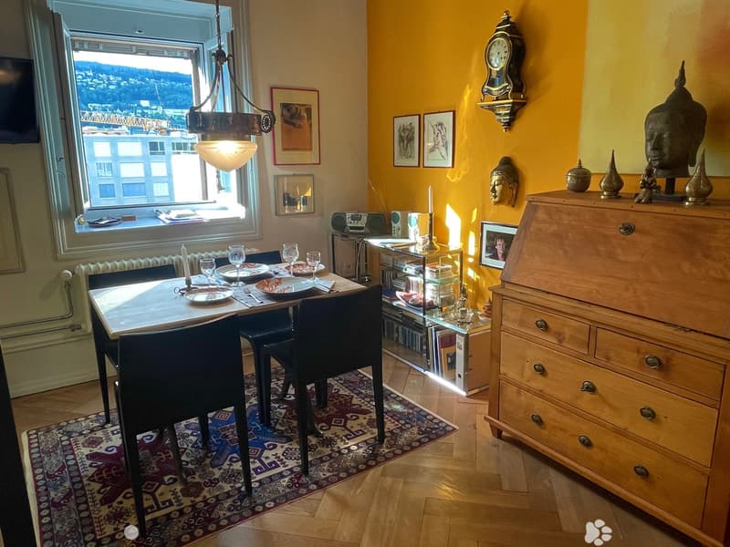 A cozy dining area with a table set for four, surrounded by chairs, a wooden cabinet, and decorative items including a clock and framed pictures. The room features warm yellow walls, wooden flooring, and a large window offering a scenic view.