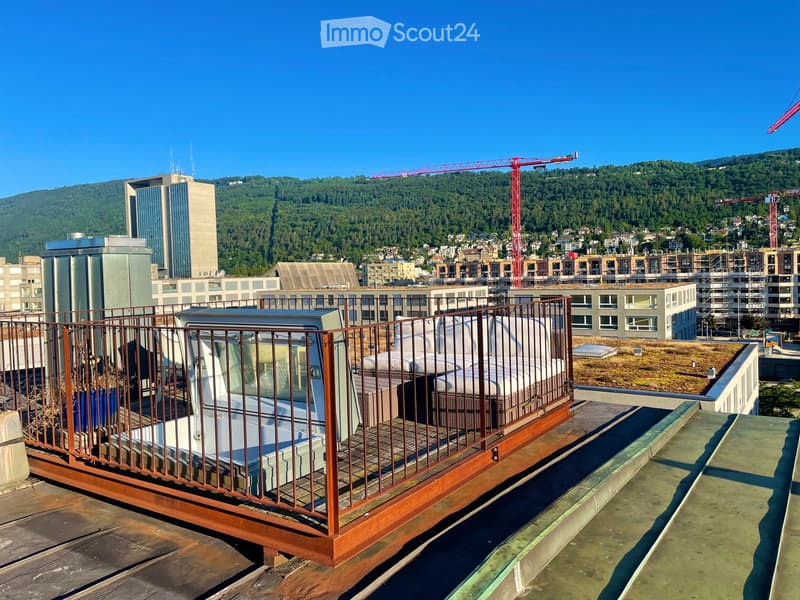 A rooftop terrace with a wooden railing, featuring a sofa and a table, overlooking a cityscape with buildings and a forested hill in the background.