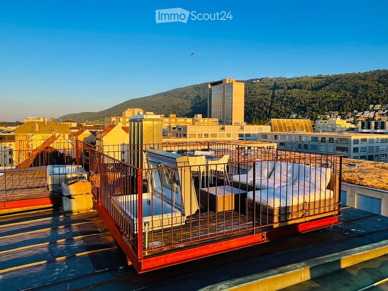 Rooftop terrace with a sofa, chairs, and a table, overlooking a cityscape with buildings and hills.