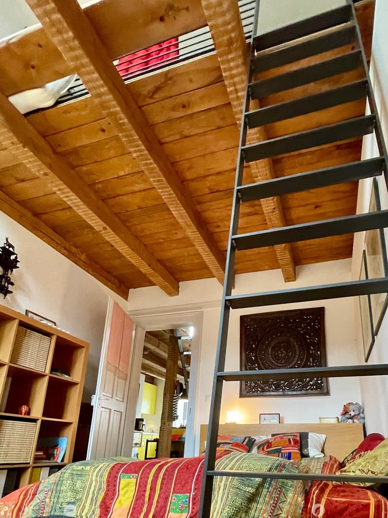 A cozy bedroom with a wooden slanted ceiling, a metal lofted bed, colorful bedding, a wooden shelf with baskets, and a mirror in the background.