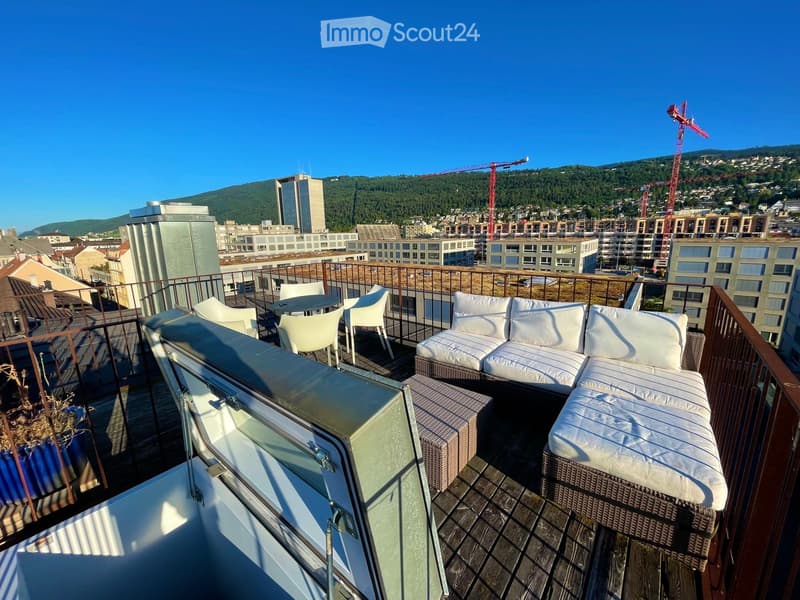 Balcony with wooden decking, white sofa, table, and chairs, overlooking a cityscape with cranes and buildings.