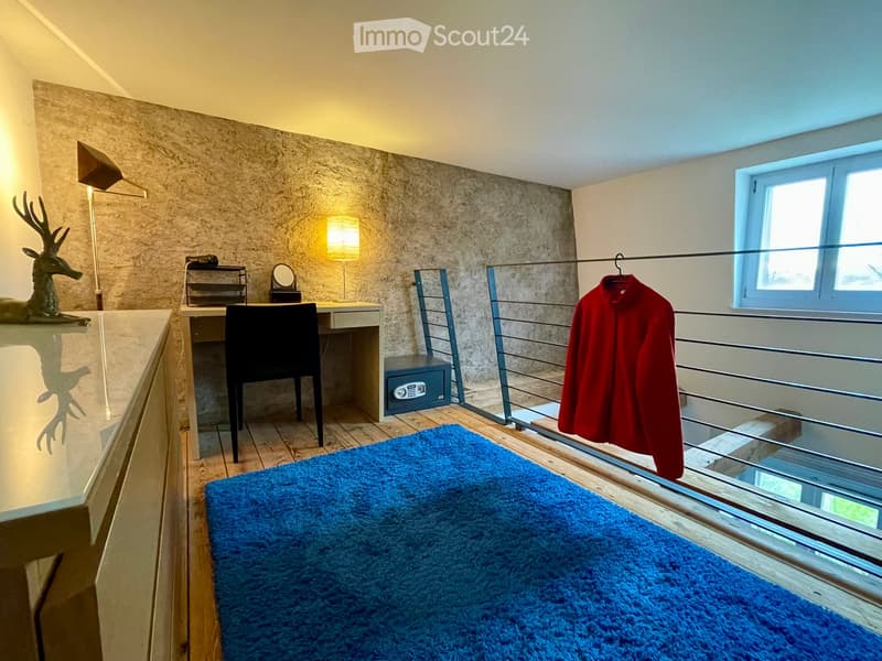 A furnished room with a desk, chair, lamp, mirror, security box, a red coat hanging on a rack, and a decorative deer statue on a shelf.