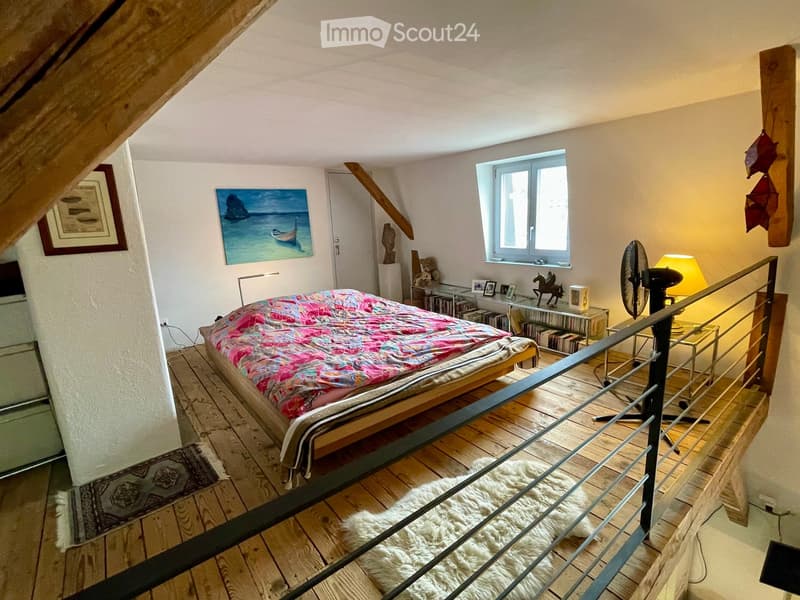 Cozy attic bedroom with wooden floors, slanted ceilings, a bed with a colorful quilt, a small table with a lamp, a fan, and decorative items including a painting and a sculpture.