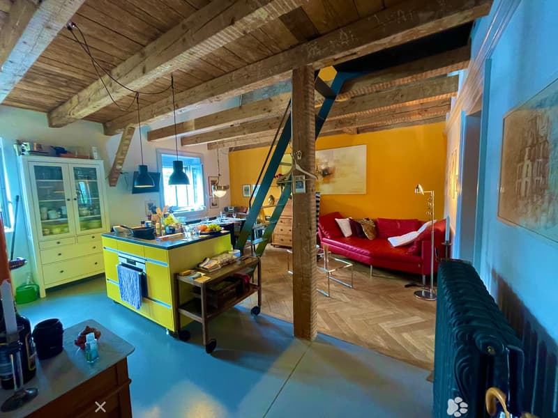 Open-plan living and dining area with exposed wooden beams, a red leather sofa, a glass coffee table, a yellow kitchen island, and pendant lights.