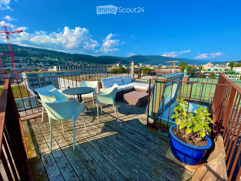 A wooden balcony with white furniture including a sofa, chairs, a small table, and a potted plant, overlooking a cityscape with mountains in the background.