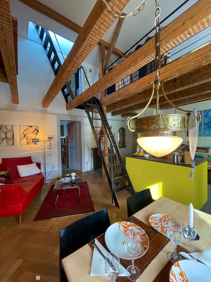 Open-plan living and dining area with exposed wooden beams, a red sofa, a glass coffee table, a dining table set for two, and a bright yellow kitchen area.