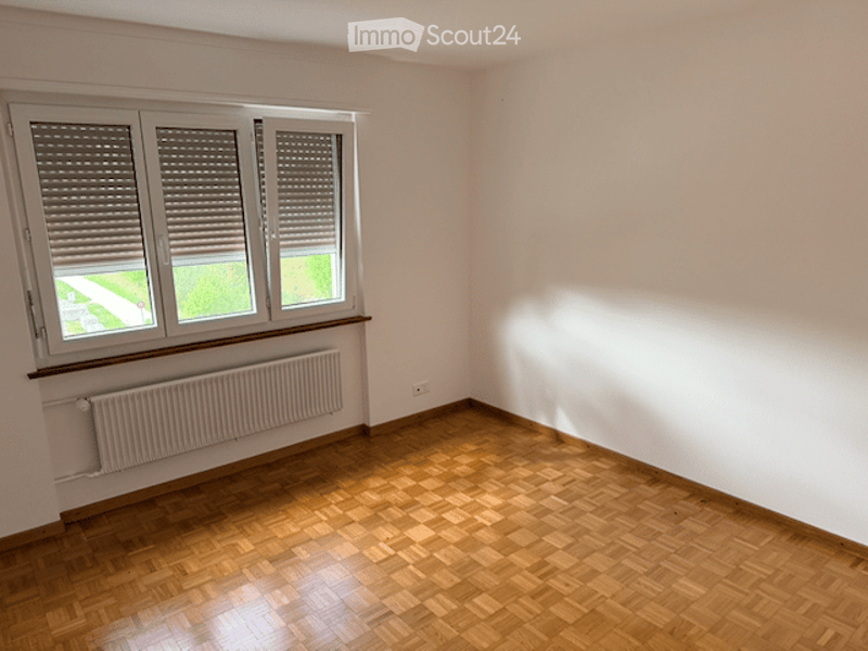 Empty room with wooden flooring, white walls, and a large window with blinds. A radiator is visible below the window.