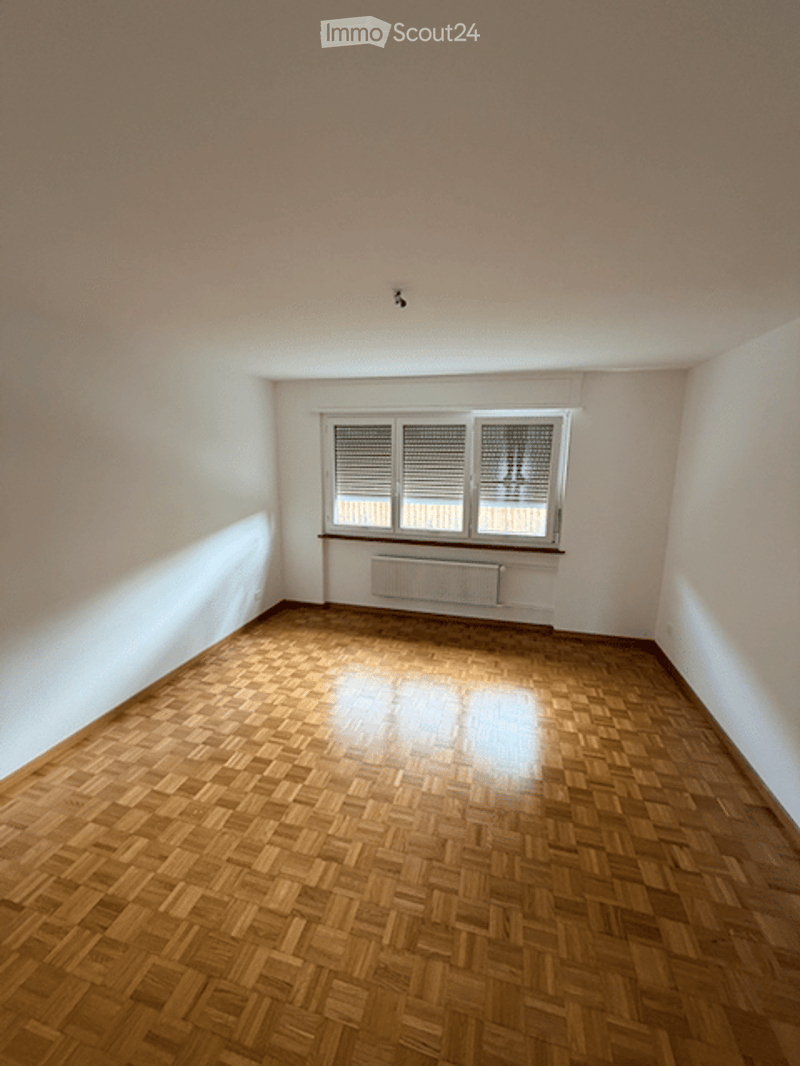 Empty room with wooden parquet flooring, white walls, and a single window with blinds. A radiator is visible below the window.