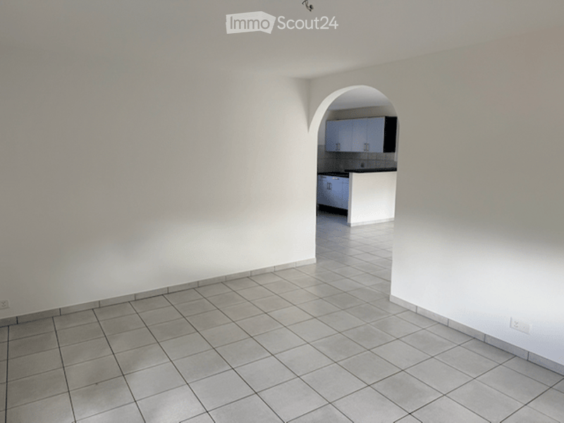 Empty room with tiled floor and white walls, featuring an arched doorway leading to a small kitchen area with cabinets and a countertop.