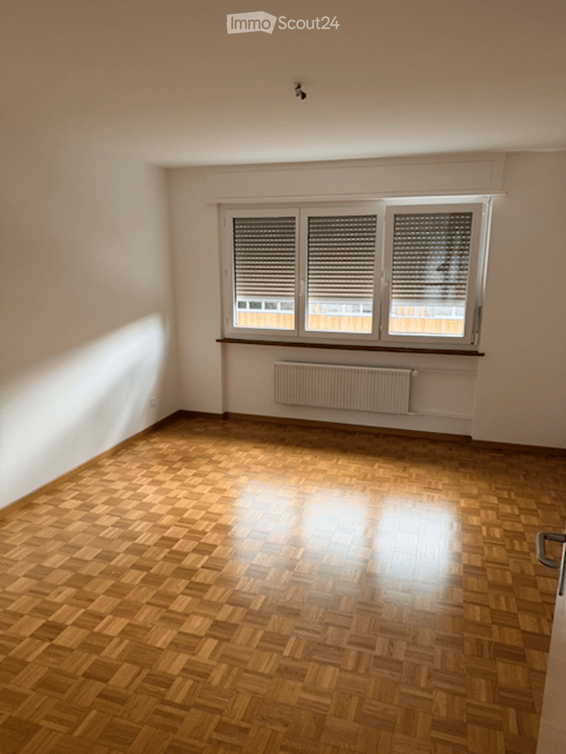 Empty room with wooden parquet flooring, white walls, and a large window with blinds. A radiator is mounted below the window.