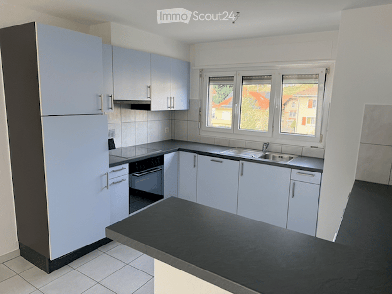 Modern kitchen with light blue cabinets, black countertops, stainless steel appliances, and large windows overlooking a residential area.